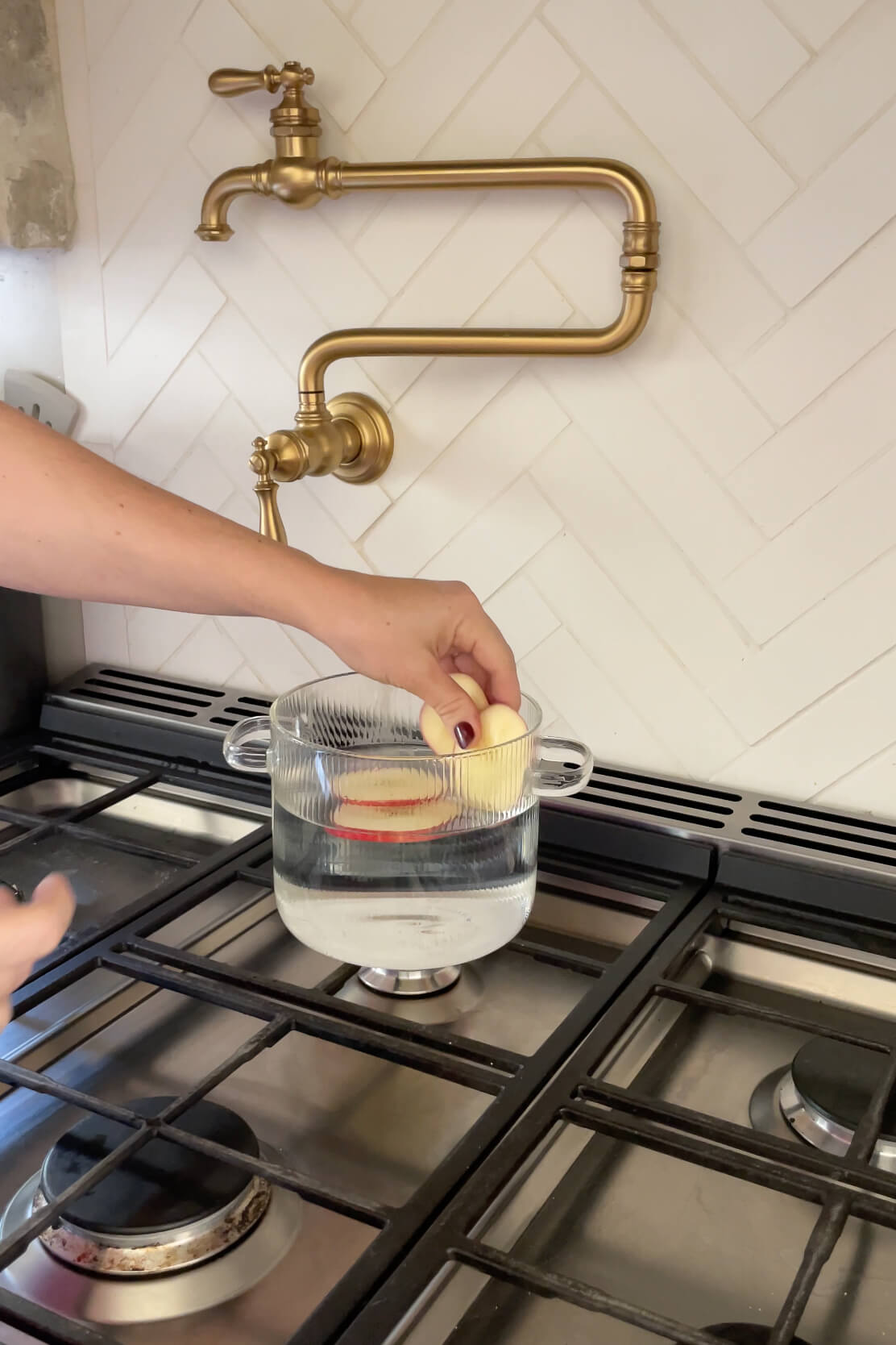 Hand placing apple slices into a glass pot filled with water on a stove beneath a brass pot filler.